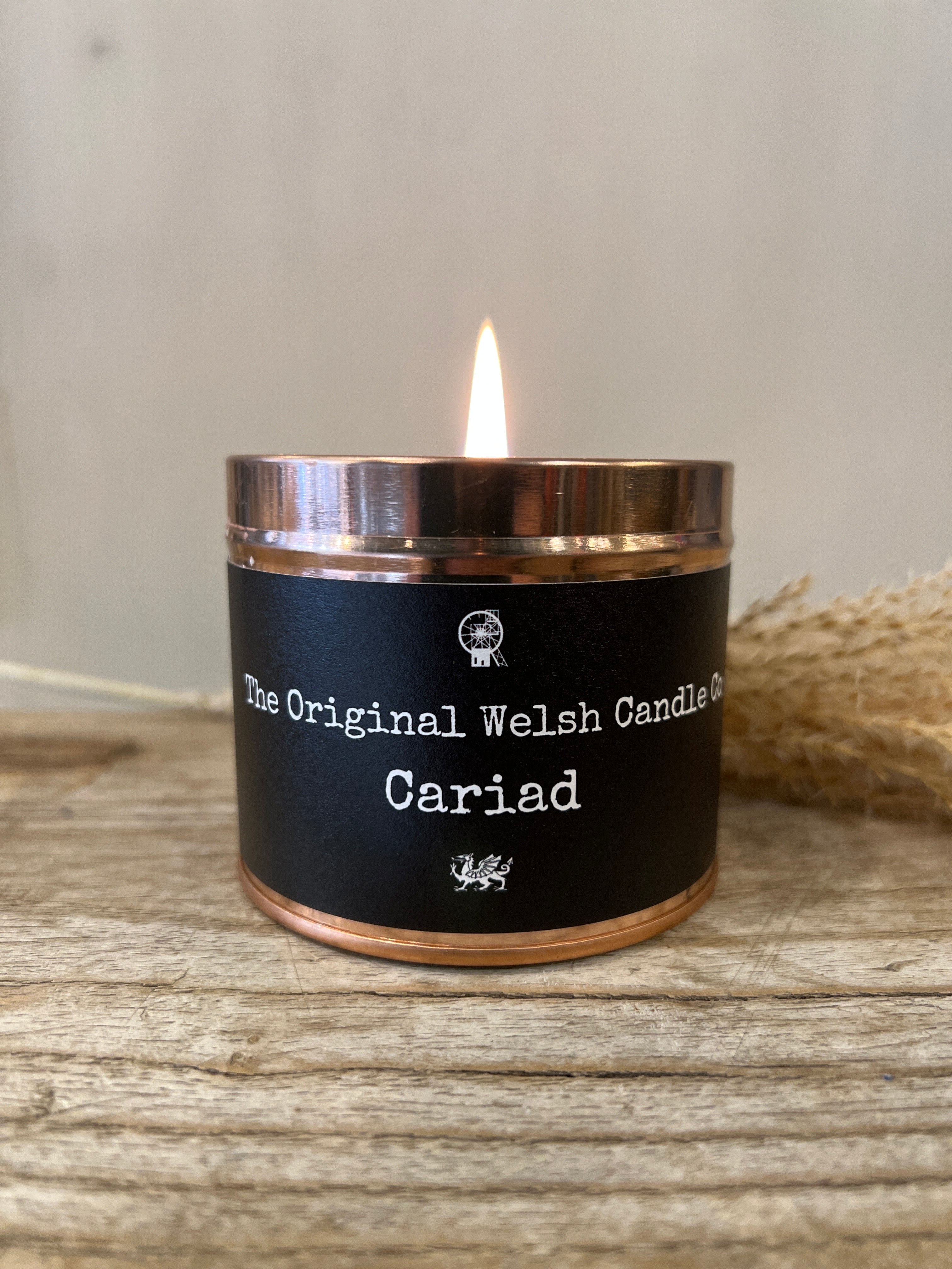 A rose gold tin scented candle with a lit flame, labeled 'The Original Welsh Candle CARIAD'.